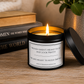 A black metal candle jar with a white label reading 'WOMEN AREN'T MEANT TO SIT AND LOOK PRETTY WE ARE MEANT TO BUILD EMPIRES' sits lit on a wooden table with its lid leaning beside it. Books and a potted plant are blurred in the background.