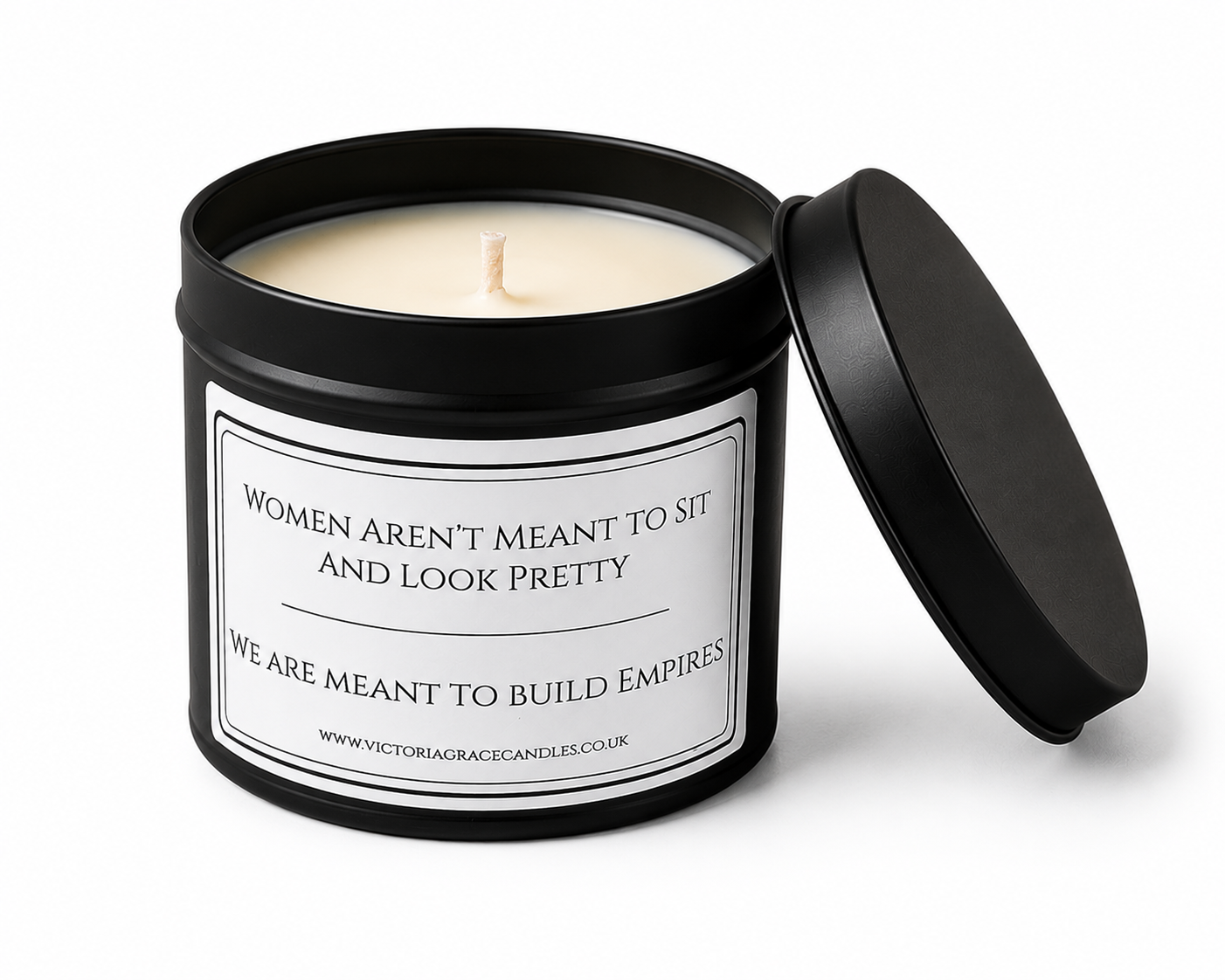 An open black metal candle tin with cream-colored wax and a lid leaning against it on a white background. The front label reads, 'WOMEN AREN'T MEANT TO SIT AND LOOK PRETTY WE ARE MEANT TO BUILD EMPIRES' with the brand Victoria Grace Candles visible at the bottom.