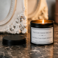 A black metal candle tin with a lit flame sits on a dark marble surface, with its black lid placed beside it. The front label reads, 'YOU CAN HAVE EXCUSES OR YOU CAN HAVE RESULTS' 
