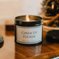 black tin candle with the words cheer up fucker in it set on a wooden sideboard. 